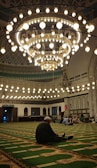The mosque's prayer hall filled with worshippers during tarawih prayers, illuminated by warm chandeliers.