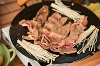 Slices of meat cooking on a black pan, surrounded by clusters of enoki mushrooms. The scene is set in a kitchen environment with a wooden spatula resting nearby.