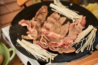 Slices of meat cooking on a black pan, surrounded by clusters of enoki mushrooms. The scene is set in a kitchen environment with a wooden spatula resting nearby.