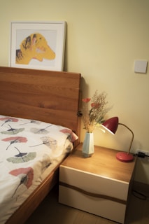 A cozy bedroom setting featuring a wooden bed with a patterned duvet cover. A framed artwork of a yellow and orange dog hangs above the bed. Next to the bed, there is a wooden nightstand with a modern red desk lamp and a geometric vase containing dried flowers. The wall is painted a soft yellow.