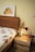 A cozy bedroom setting featuring a wooden bed with a patterned duvet cover. A framed artwork of a yellow and orange dog hangs above the bed. Next to the bed, there is a wooden nightstand with a modern red desk lamp and a geometric vase containing dried flowers. The wall is painted a soft yellow.