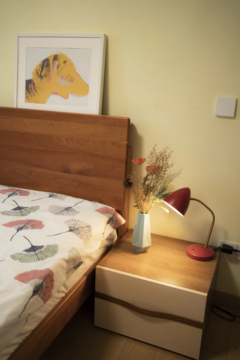 A cozy bedroom setting featuring a wooden bed with a patterned duvet cover. A framed artwork of a yellow and orange dog hangs above the bed. Next to the bed, there is a wooden nightstand with a modern red desk lamp and a geometric vase containing dried flowers. The wall is painted a soft yellow.