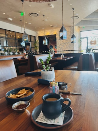 A cozy restaurant interior with modern decor. The foreground features a wooden table set with a black mug on a saucer, a small dish of seasoning, a bowl of tortilla chips, and a plant in a striped pot. The background shows brown leather seating, hanging lanterns, and large windows letting in natural light.