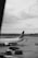 A black and white image of an airport runway featuring a parked commercial airplane with the words Garuda Indonesia visible on its fuselage. The tarmac shows several cargo pallets covered in netting. A lone person walks near the aircraft, and the background includes a cloudy sky and distant airport buildings.