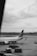 A black and white image of an airport runway featuring a parked commercial airplane with the words Garuda Indonesia visible on its fuselage. The tarmac shows several cargo pallets covered in netting. A lone person walks near the aircraft, and the background includes a cloudy sky and distant airport buildings.