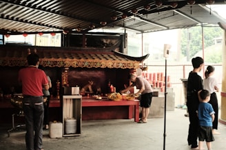 Several people are standing and interacting in an open area of a temple-like structure. The area contains a covered shrine with ornate decorations and traditional red accents. A man is placing offerings on the altar, while others seem to be observing or waiting their turn. The space is filled with natural light coming through open sides, and there are red cylindrical objects stacked in the background.