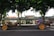 Two horse-drawn carriages pass by each other on a road lined with lush green trees. Behind them stands a building with a sign that reads 'Bali Hotel,' featuring potted plants and a large entrance.