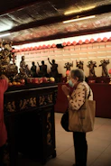 An elderly woman wearing a mask and carrying a bag stands inside a temple, hands pressed together in prayer. The temple is decorated with statues of deities and offerings on an ornate altar. Dim lighting with warm hues and red lantern decorations create a solemn and respectful atmosphere.