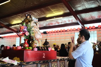 A man is holding incense while facing a statue of a smiling, rotund figure holding an ingot. The statue is adorned with flowers and other offerings, placed on a red altar with the number '2' visible. In the background, there are other people, flowers, and decorations, along with red lanterns hanging under a metallic ceiling.