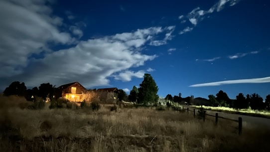 A serene night scene featuring a rustic cabin warmly lit from within. It is surrounded by trees and grassy terrain. The sky above is a deep blue, adorned with scattered clouds and a hint of stars visible in the clear sections.