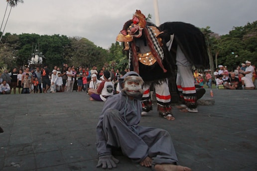 A street performance is taking place with performers in traditional costumes, including a person dressed in gray with a striking mask and another in a large, elaborate costume with gold and red decorations. A crowd of people stands in the background, watching the performance, with trees and a cloudy sky visible.