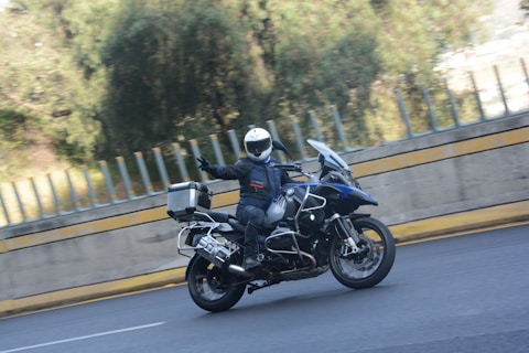 A motorcyclist wearing protective gear, including a helmet and gloves, rides a large touring motorcycle with panniers along a road with a concrete barrier. The motorcyclist is making a gesture with one hand while riding through a slightly blurred background of greenery.