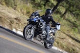 A motorcyclist dressed in black gear rides a blue adventure motorcycle on a winding road through a forested area. The rider wears a full-face helmet and the bike is equipped with side panniers and additional features for long-distance travel.