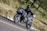 A motorcyclist dressed in black gear rides a blue adventure motorcycle on a winding road through a forested area. The rider wears a full-face helmet and the bike is equipped with side panniers and additional features for long-distance travel.
