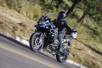 A motorcyclist dressed in black gear rides a blue adventure motorcycle on a winding road through a forested area. The rider wears a full-face helmet and the bike is equipped with side panniers and additional features for long-distance travel.