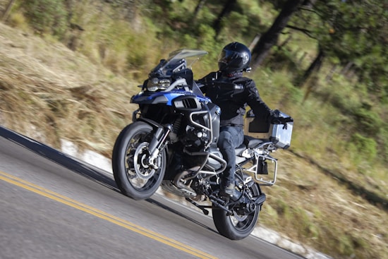 A motorcyclist dressed in black gear rides a blue adventure motorcycle on a winding road through a forested area. The rider wears a full-face helmet and the bike is equipped with side panniers and additional features for long-distance travel.