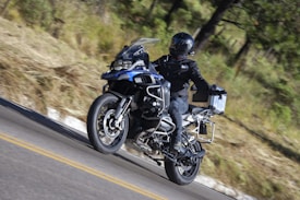 A motorcyclist dressed in black gear rides a blue adventure motorcycle on a winding road through a forested area. The rider wears a full-face helmet and the bike is equipped with side panniers and additional features for long-distance travel.