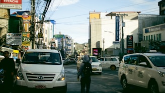 A busy urban street with several vehicles, including white vans and taxis, in motion or stopped. There are numerous commercial buildings on both sides, with signage for banks and various businesses, among them Metrobank and Caltex. Many wires crisscross above the street, and a few pedestrians are visible walking amidst the traffic.