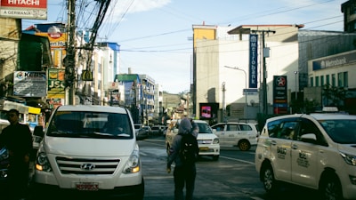 A busy urban street with several vehicles, including white vans and taxis, in motion or stopped. There are numerous commercial buildings on both sides, with signage for banks and various businesses, among them Metrobank and Caltex. Many wires crisscross above the street, and a few pedestrians are visible walking amidst the traffic.