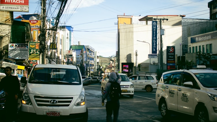 A busy urban street with several vehicles, including white vans and taxis, in motion or stopped. There are numerous commercial buildings on both sides, with signage for banks and various businesses, among them Metrobank and Caltex. Many wires crisscross above the street, and a few pedestrians are visible walking amidst the traffic.