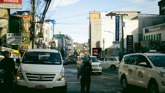 A busy urban street with several vehicles, including white vans and taxis, in motion or stopped. There are numerous commercial buildings on both sides, with signage for banks and various businesses, among them Metrobank and Caltex. Many wires crisscross above the street, and a few pedestrians are visible walking amidst the traffic.