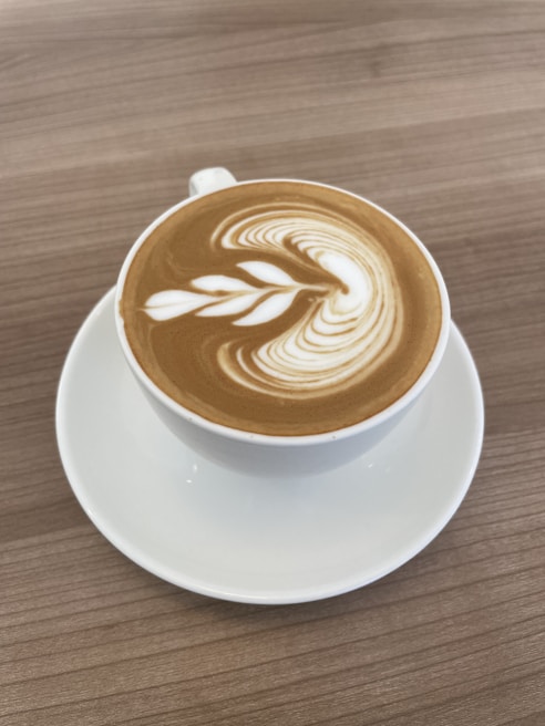 A cup of coffee with a beautiful leaf-shaped latte art on top, placed on a white saucer. The coffee has a creamy surface with intricate patterns, set on a wooden table.