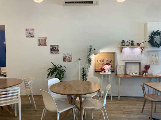 A cozy caf&eacute; interior featuring wooden tables and white chairs. Decorative elements include wall posters, potted plants, and a festive atmosphere with holiday decorations such as wreaths and a small Santa doll. A wooden shelf holds additional decorative items. The caf&eacute; exhibits a clean and minimalist design.