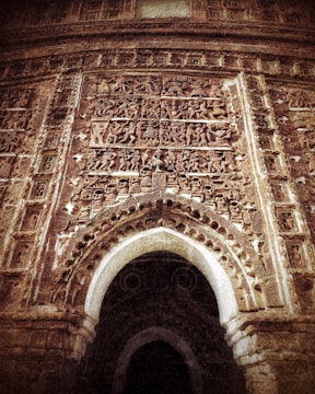 An intricately carved stone facade of an ancient structure, featuring detailed relief sculptures depicting figures and scenes. The arched doorway is prominently framed with ornate designs, showcasing artistry and cultural heritage. The weathered surface suggests historical significance and antiquity.