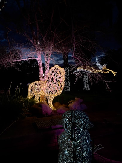 A nighttime scene featuring illuminated sculptures, including a large, glowing lion and a figure that seems to be a flying bird or angel. The sculptures are formed from intricate light patterns, casting a warm, colorful glow against the dark sky and bare trees in the background.