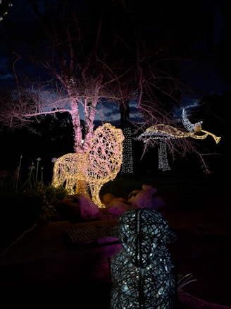 A nighttime scene featuring illuminated sculptures, including a large, glowing lion and a figure that seems to be a flying bird or angel. The sculptures are formed from intricate light patterns, casting a warm, colorful glow against the dark sky and bare trees in the background.