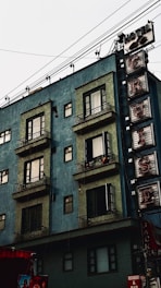 A multi-story building with a greenish-blue facade features several windows with elaborate iron railings. A vertically-oriented sign with the word 'CRUISE' in large letters is attached to the building. Overhead, numerous electrical wires crisscross. The lower part of the building includes a red awning and several advertisements.