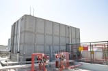 A large industrial water storage tank is situated on a rooftop, surrounded by various pipes, valves, and safety equipment. There is a metal ladder attached to the tank. A cage-like fence with warning signs encloses part of the area.