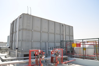 A large industrial water storage tank is situated on a rooftop, surrounded by various pipes, valves, and safety equipment. There is a metal ladder attached to the tank. A cage-like fence with warning signs encloses part of the area.