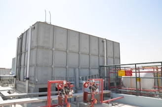 A large industrial water storage tank is situated on a rooftop, surrounded by various pipes, valves, and safety equipment. There is a metal ladder attached to the tank. A cage-like fence with warning signs encloses part of the area.