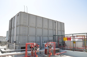 A large industrial water storage tank is situated on a rooftop, surrounded by various pipes, valves, and safety equipment. There is a metal ladder attached to the tank. A cage-like fence with warning signs encloses part of the area.