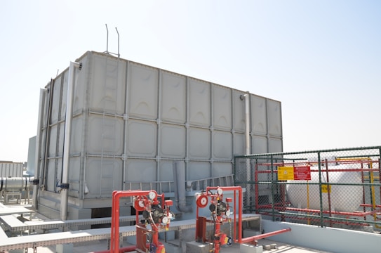 A large industrial water storage tank is situated on a rooftop, surrounded by various pipes, valves, and safety equipment. There is a metal ladder attached to the tank. A cage-like fence with warning signs encloses part of the area.