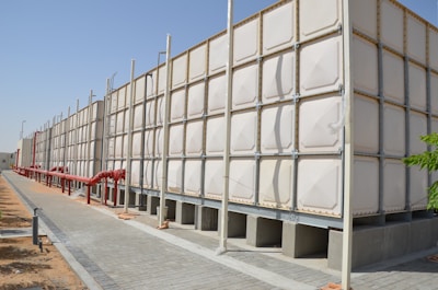 A large industrial water tank with sections made of a light-colored material, supported by a metal frame and beams. Red pipes run alongside the tank, attached to the ground. The setting is outdoors under a clear blue sky, with a paved walkway and some patches of sandy soil visible.