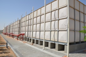 A large industrial water tank with sections made of a light-colored material, supported by a metal frame and beams. Red pipes run alongside the tank, attached to the ground. The setting is outdoors under a clear blue sky, with a paved walkway and some patches of sandy soil visible.