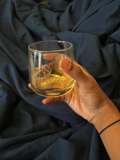 A hand holding a glass filled with a yellow liquid, set against a dark blue fabric backdrop. The glass has some text etched on it, and the hand is adorned with a thin black bracelet.