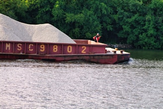 A barge loaded with gravel is floating on a river, accompanied by lush green trees in the background. A person wearing a life jacket is standing on the deck of the barge.