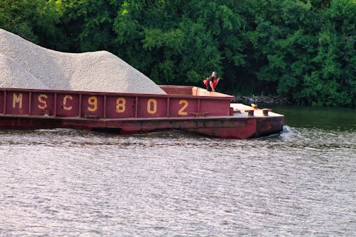 A barge loaded with gravel is floating on a river, accompanied by lush green trees in the background. A person wearing a life jacket is standing on the deck of the barge.
