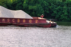 A barge loaded with gravel is floating on a river, accompanied by lush green trees in the background. A person wearing a life jacket is standing on the deck of the barge.