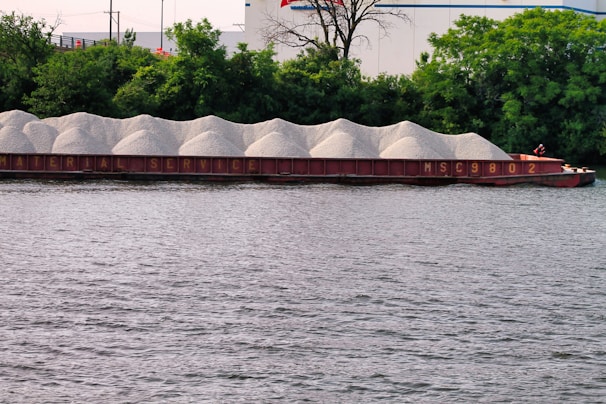 A barge loaded with piles of gravel is floating on a river, with a backdrop of dense green trees and some industrial buildings. The barge is labeled with the words 'Material Service' and the number 'MSC9802'.