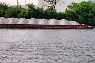 A barge loaded with piles of gravel is floating on a river, with a backdrop of dense green trees and some industrial buildings. The barge is labeled with the words 'Material Service' and the number 'MSC9802'.