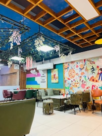 The image depicts a vibrant and colorful cafe or restaurant interior. The ceiling is styled with a wooden lattice design featuring hanging plants and a painted sky backdrop. Walls are adorned with playful and artistic murals, featuring various food items, including pizza. Multiple tables and chairs are arranged throughout, with a mix of materials like upholstered green chairs and wicker-style stools. A wall sign humorously reads, 'I'm sorry for what I said when I was hungry.'