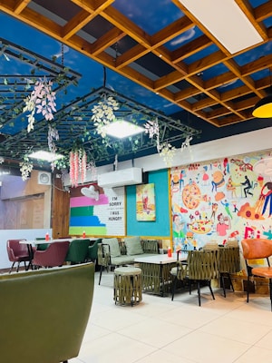 The image depicts a vibrant and colorful cafe or restaurant interior. The ceiling is styled with a wooden lattice design featuring hanging plants and a painted sky backdrop. Walls are adorned with playful and artistic murals, featuring various food items, including pizza. Multiple tables and chairs are arranged throughout, with a mix of materials like upholstered green chairs and wicker-style stools. A wall sign humorously reads, 'I'm sorry for what I said when I was hungry.'