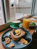 A cozy café setting featuring a wooden table with a delicious breakfast spread. The plate contains French toast topped with powdered sugar, berries, nuts, and a dollop of butter. Next to it, a bright orange juice and a green cup of cappuccino with latte art are placed. Through the window, a blurred view of the street is visible.