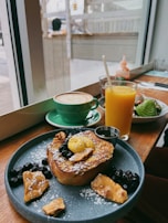 A cozy café setting featuring a wooden table with a delicious breakfast spread. The plate contains French toast topped with powdered sugar, berries, nuts, and a dollop of butter. Next to it, a bright orange juice and a green cup of cappuccino with latte art are placed. Through the window, a blurred view of the street is visible.