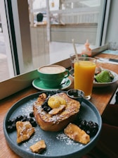 A cozy café setting featuring a wooden table with a delicious breakfast spread. The plate contains French toast topped with powdered sugar, berries, nuts, and a dollop of butter. Next to it, a bright orange juice and a green cup of cappuccino with latte art are placed. Through the window, a blurred view of the street is visible.