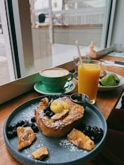 A cozy café setting featuring a wooden table with a delicious breakfast spread. The plate contains French toast topped with powdered sugar, berries, nuts, and a dollop of butter. Next to it, a bright orange juice and a green cup of cappuccino with latte art are placed. Through the window, a blurred view of the street is visible.